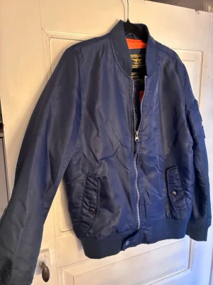 Mörkblå bomberjacka från Alpha Industries - Snygg mörkblå bomberjacka från Alpha Industries med klassisk orange insida. Jackan har dragkedja framtill, två fickor och en innerficka med knappstängning, en ficka på vänster ärm med dragkedja och ribbade muddar vid ärmslut och nederkant. Tillverkad i slitstarkt syntetmaterial och har en cool, tidlös look.