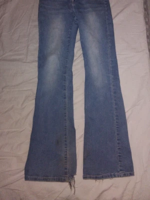 Blå bootcut jeans med detaljer - Säljer ett par ljusblå bootcut jeans med slitningar och snygga broderade bakfickor med vita knappar. Jeansen har klassisk femficksmodell och är tillverkade i denim. Perfekta för dig som gillar retrostil och vill ha ett par jeans med unik touch.