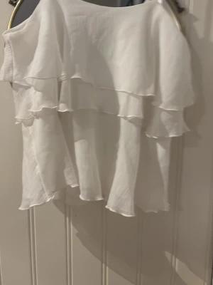 Vit volang topp med offshoulder - Säljer en superfin vit topp med dubbla volanger och offshoulder-design. Toppen har en luftig och flowy känsla, perfekt för varma dagar. Materialet känns lätt och svalt, och volangerna ger en romantisk vibe.