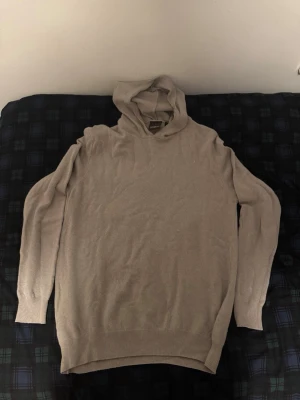 Beige stickad hoodie Oscar Jacobson - Snygg beige stickad hoodie från Oscar Jacobson. Tillverkad i Italien med mjukt material och ribbade muddar vid ärmslut och nederkant. Klassisk huva och lång ärm för en avslappnad men stilren look.