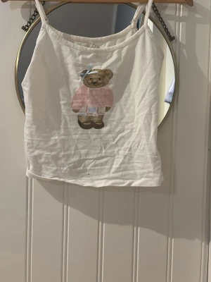 Vit linnetopp med nalletryck - Söt vit linnetopp med tunna axelband och ett tryck av en nallebjörn i rosa tröja och blå rosett framtill. Perfekt för varma dagar och enkel att matcha med jeans eller kjol.