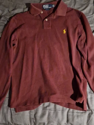 Vinröd långärmad piké från Polo Ralph Lauren - Säljer en vinröd långärmad pikétröja från Polo Ralph Lauren med klassisk krage och två knappar. Tröjan har det ikoniska gula broderade logotypen på bröstet