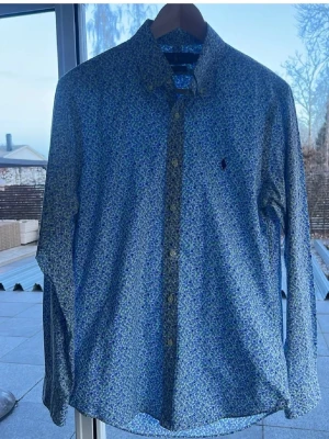 Blommig skjorta från Ralph Lauren - Snygg slim fit skjorta från Ralph Lauren med blått och grönt blommönster. Skjortan har långa ärmar, klassisk krage och knappar framtill. Tillverkad i 100% bomull med stretch för extra komfort. Perfekt för dig som gillar stilrena detaljer och mönster.