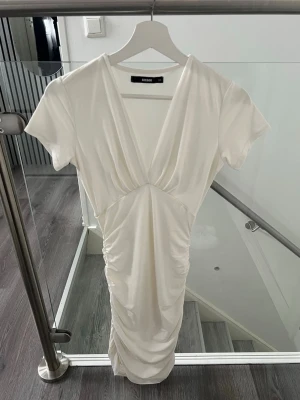 Studentklänning från BikBok, XS - Säljer en vit klänning från BikBok med  v-ringning. Klänningen har korta ärmar och snygg rynkning längs sidorna. Passar perfekt som en studentklänning💖
