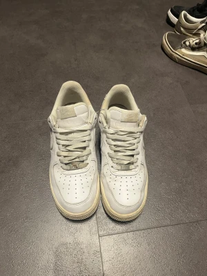 Vita Nike Air Force 1 sneakers - Klassiska vita Nike Air Force 1 sneakers i lågt utförande. Skorna har en vit ovandel i läder med perforeringar på tån och den ikoniska swoosh-loggan på sidan. Yttersulan är något gulnad vilket ger en retrovibe. Perfekta för dig som gillar streetwear och tidlös stil.