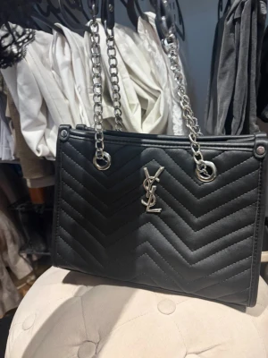 Svart YSL quiltad handväska - Snygg svart quiltad handväska från YSL med silverfärgade kedjehandtag och ikonisk YSL-logga framtill. Väskan har ett stilrent mönster i chevron-design och är tillverkad i ett slätt material som ger en lyxig känsla. Perfekt accessoar för att lyfta din outfit.