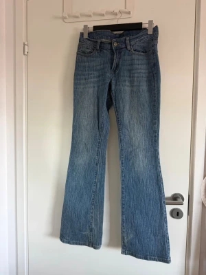 Blå bootcut jeans med vintagekänsla - Säljer ett par blå bootcut jeans med klassisk femficksmodell och ljusa slitningar framtill. Jeansen har en något utsvängd passform nedtill och är tillverkade i denim med tydlig struktur. Perfekta för dig som gillar retrostil och vill ha ett par bekväma jeans med snygg tvätt.