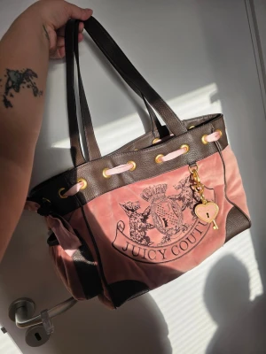 Juicy Couture daydreamer väska - Juicy Couture handväska i rosa sammet med bruna detaljer i konstläder. Väskan har guldiga metalldetaljer, ett emblem framtill, rosa band och en dekorativ scarf på sidan. Hängande hjärta och nyckel-charm ingår. Rymlig modell med dubbla axelband.