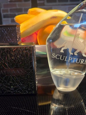 Sculpture och Ryhaan Terra  - Två parfymflaskor: en genomskinlig Sculpture-flaska med elegant lejonmotiv och vit text, samt en mörk, mönstrad Ayhaan-flaska med metallisk finish. Båda flaskorna har unika former och detaljer som gör dem till snygga inslag på parfymhyllan.