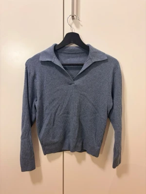 Blå ribbstickad tröja med krage - Snygg blå ribbstickad tröja med klassisk krage och v-ringning. Tröjan har långa ärmar och är gjord i ett mjukt, stretchigt material som sitter skönt på kroppen. Perfekt för dig som gillar en stilren och avslappnad look.