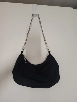 Svart axelväska med kedjerem - Trendig svart handväska i mjukt tyg med silverfärgad kedjerem och dragkedja upptill. Väskan har en rundad form och minimalistisk design som passar till många olika stilar. Perfekt för dig som gillar en enkel men cool look.