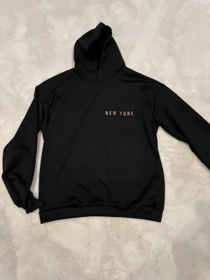 Svart hoodie med New York-tryck - Snygg svart hoodie med huva och texten 'NEW YORK' tryckt i vitt på bröstet. Hoodien har en klassisk passform med långa ärmar och ribbade muddar. Perfekt för dig som gillar enkel streetstyle och vill ha en stilren look.