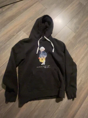 Svart hoodie Polo Ralph Lauren M - Svart hoodie från Polo Ralph Lauren med klassisk björn-illustration på bröstet. Hoodien har vit dragsko i huvan och en stor ficka framtill. Tillverkad i mjukt material som känns skönt mot huden. Perfekt för en avslappnad och stilren look.