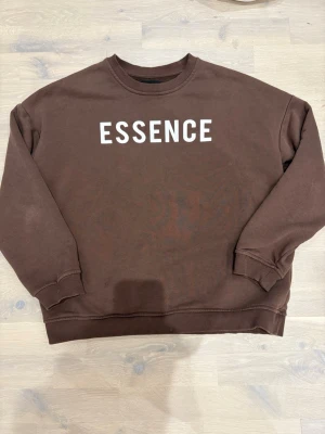 Brun sweatshirt med ESSENCE-tryck - Cool brun sweatshirt med vit text 'ESSENCE' på bröstet. Tröjan har rund hals, ribbade muddar vid ärmslut och nederkant samt en avslappnad passform. Perfekt för en casual streetwear-look.