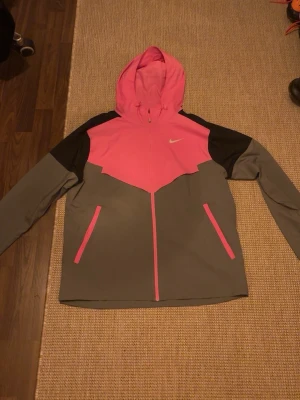 Nike vindjacka rosa och grå - Snygg vindjacka från Nike i grått och rosa med svarta axelpartier. Jackan har huva, hel dragkedja framtill och två fickor med rosa detaljer. Materialet är lätt och vindtätt, perfekt för blåsiga dagar. Nike-logga på bröstet.