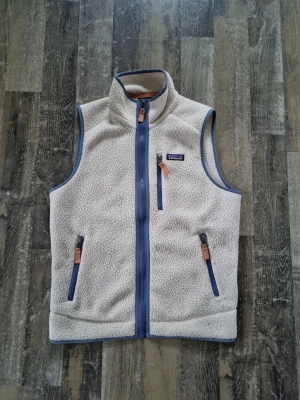 Beige fleeceväst från Patagonia - Mysig beige fleeceväst från Patagonia med blå detaljer och dragkedjor. Västen har hög krage, tre fickor med dragkedja och snygga läderdetaljer på dragkedjorna. Perfekt lager-på-lager-plagg för kyliga dagar.