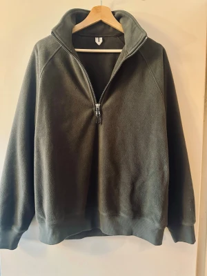 Mörkgrön fleece hoodie med halv dragkedja - Mysig mörkgrön hoodie i fleece med halv dragkedja framtill och hög krage. Tröjan har ribbade muddar vid ärmslut och nederkant. Perfekt för lager på lager och chill dagar. Enkel och stilren design utan synligt märke.