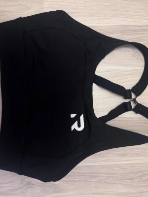 Svart sport-bh från Relode - Säljer en svart sport-bh från Relode med brottarrygg och justerbara axelband. Bh:n har en bred resår under bysten och ett vitt Relode-logotyptryck framtill. Materialet är stretchigt och passar perfekt för träning.