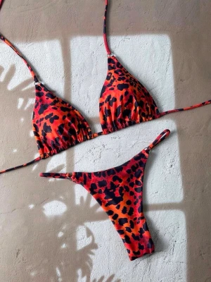 Leopardmönstrad bikini i rött och svart - Trendig bikini med leopardmönster i rött, orange och svart. Överdelen har trekantskupor med smala knytband och underdelen är en klassisk tanga-modell med tunna sidoband. Perfekt för strandhäng och poolparty. Använd en gång.