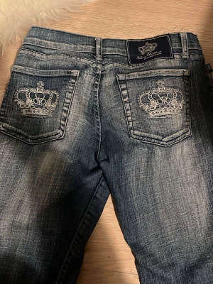 Mörkblåa jeans med kronbrodyr victoria beckham - Säljer ett par mörkblå jeans från Von Dutch med slitningar och snygga broderade kronor på bakfickorna. Jeansen har klassisk femficksdesign, raka ben och cool tvättad look. Perfekta för dig som gillar streetstyle och detaljer.