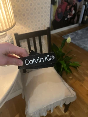 Svart Calvin Klein bälte - Snyggt svart bälte från Calvin Klein med vit logga tryckt längs hela bandet. Bältet har en enkel och modern design som passar till många olika outfits. Perfekt accessoar för dig som gillar stilrena detaljer och vill ha ett bälte som sticker ut.