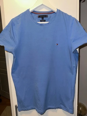 Blå t-shirt från Tommy Hilfiger - Snygg ljusblå t-shirt från Tommy Hilfiger i ekologisk bomull. Slim fit-modell med korta ärmar och klassisk rund halsringning. Liten broderad logga på bröstet ger en clean och stilren look. Priset kan diskuteras pga liten fläck, nästan helt osynligt för blotta ögat, syndligt på kamera med blixt.