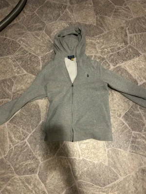 Grå zip hoodie från Polo Ralph Lauren - Säljer en grå hoodie från Polo Ralph Lauren med dragkedja och huva. Klassisk blå logga broderad på bröstet. Tillverkad i mjukt sweatshirtmaterial som är skönt mot huden. Perfekt för en avslappnad och stilren look. Använd ganska mycket därför skapligt pris