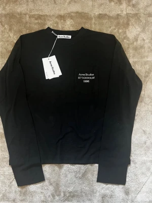 Acne Studios tröja - Svart långärmad tröja från Acne Studios med rund hals och bröstficka. På fickan finns vit broderad text med 'Acne Studios Stockholm 1996'. Tillverkad i mjuk bomull för en clean och stilren look.