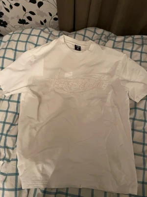 Vit t-shirt från Stussy med broderi - Säljer en vit t-shirt från Stussy med ett stort, ton-i-ton broderat Stussy-logo på bröstet. Klassisk passform och rund halsringning. Perfekt för dig som gillar streetwear och vill ha en clean look med snygga detaljer.