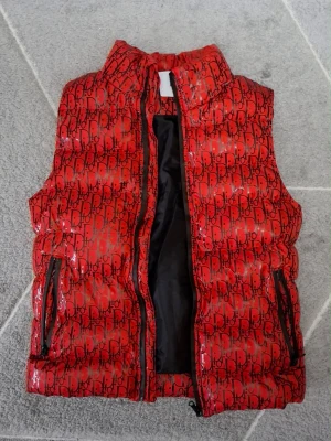 Röd dunväst från Dior med logomönster - Säljer en röd dunväst från Dior med svart logomönster över hela västen. Västen har hög krage, två dragkedjeförsedda fickor och svart innerfoder. Perfekt för dig som vill sticka ut med en exklusiv och trendig look.