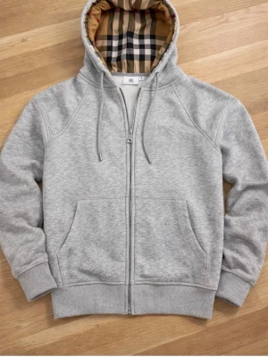 Grå zip hoodie från Burberry - Snygg grå zip hoodie från Burberry med klassiskt rutigt foder i huvan. Hoodien har dragsko i huvan, två stora fickor fram och ribbade muddar. Tillverkad i mjuk bomullsblandning för extra komfort. Perfekt för en avslappnad och stilren look.
