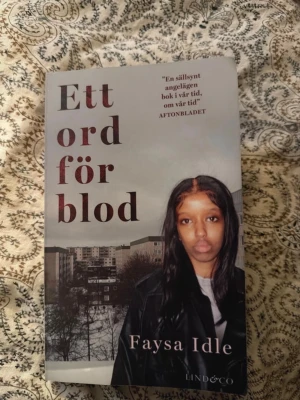 Ett ord för blod av Faysa Idle - Romanen 'Ett ord för blod' av Faysa Idle är en pocketbok med omslag i grå och mörka toner. Omslaget visar en person framför ett höghuslandskap. Boken utforskar livet i Stockholms förorter och gängvåldets påverkan på unga kvinnor.