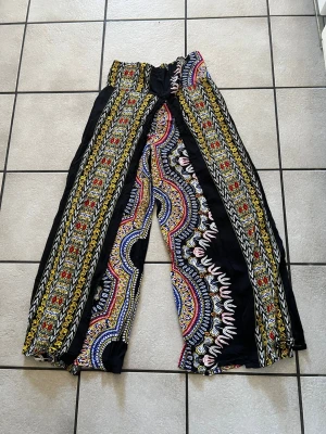 Färgglada vida byxor med mönster storlek S/M - Säljer ett par riktigt snygga byxor med vida ben och elastisk midja. De är svarta i grunden och har färgstarka etnoinspirerade mönster i gult, rött, blått och vitt. Perfekta för dig som vill sticka ut och ha något bekvämt och luftigt. Materialet känns lätt och mjukt. Midja 70 cm, bomull 