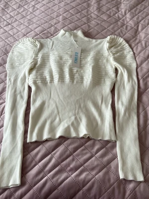 Vit ribbad topp från Blue Vanilla - Trendig vit ribbad topp från Blue Vanilla med puffade axlar och hög krage. Toppen har lång ärm och är tillverkad i ett stretchigt material som sitter snyggt på kroppen. Perfekt att styla med jeans eller kjol för en cool look.
