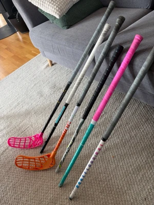 Flera innebandyklubbor i olika färger - Säljer ett gäng innebandyklubbor i olika färger och modeller. Klubborna har blad i rosa och orange samt skaft i svart, vitt, turkost och grått. Några har färgglada detaljer och grepptejp i rosa och svart. Perfekt för dig som vill ha flera alternativ till träning eller match. En icon klubba från Salming 27 flex so. Är 90 cm lång. Sen en trasig hyperligtht som har gått av längst ner som är 88 cm lång. Sen lite andra klubbor som är mellan 70 till 85 cm långa