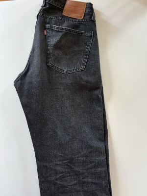 Svarta Levi's 501 jeans straight fit - Säljer ett par svarta Levi's 501 jeans med klassisk straight fit och femficksmodell. Jeansen har en vintage look med tydliga sömmar, läderpatch bak i midjan och röd Levi's-tag på bakfickan. Tillverkade i slitstark bomullsdenim.