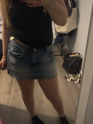 Blå jeanskjol med slitningar - Säljer en snygg blå jeanskjol i kort modell med slitna detaljer framtill. Kjolen har klassiska fickor både fram och bak samt knapp och dragkedja i midjan. Perfekt för dig som gillar en avslappnad och trendig stil.