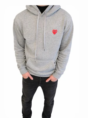 Comme des Garçons Play - Grå hoodie från Comme des Garçons Play med klassisk huva och dragsnören. På bröstet sitter det ikoniska röda hjärtat med ögon. Hoodien har en stor magficka och är tillverkad i mjuk bomullsblandning. Perfekt för en avslappnad streetwear-look.