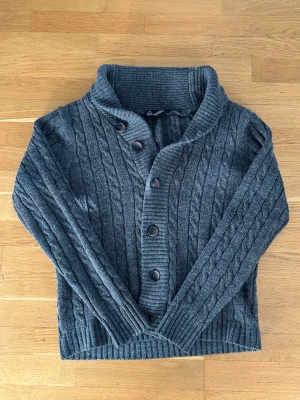 Mörkgrå kabelstickad kofta - Skön mörkgrå kabelstickad kofta med stora knappar framtill och bred krage. Koftan har långa ärmar och ribbade muddar vid ärmslut och nederkant. Perfekt för lager på lager och en chill look.