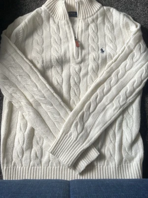 Vit kabelstickad tröja Polo Ralph Lauren - Kabelstickad vit tröja från Polo Ralph Lauren med halv dragkedja och ribbade muddar. Klassisk marinblå logga broderad på bröstet. Tröjan har hög krage och är gjord i mjukt material, perfekt för kyliga dagar.