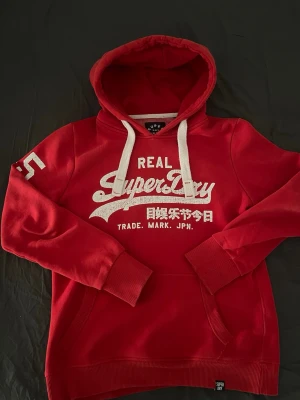 Röd hoodie från Superdry - Snygg röd hoodie från Superdry med vit logga och text på bröstet samt tryck på ena ärmen. Hoodien har vita dragsnören och en klassisk känguruficka framtill. Mjuk insida och ribbade muddar vid ärmar och nederkant. Perfekt för en avslappnad streetstyle.