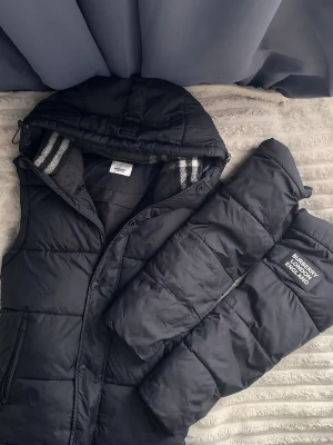 Svart jacka  från Burberry - Passar också för size S. Snygg svart dunväst från Burberry med avtagbar huva och quiltad design. Västen har en patch med Burberry-logga på ena sidan och dragkedja framtill. Insidan har randiga detaljer vid kragen. Perfekt för lager-på-lager och streetwear.