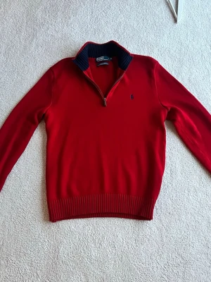 Röd stickad tröja från Polo Ralph Lauren - Snygg röd stickad tröja från Polo Ralph Lauren med halv dragkedja och mörkblå krage. Tröjan har långa ärmar, ribbade muddar och den klassiska broderade loggan på bröstet. Perfekt för dig som gillar stilrena och tidlösa plagg.