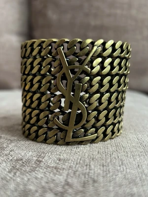 Brett kedjearmband från Yves Saint Laurent - Coolt och brett armband i antikguld från Yves Saint Laurent. Armbandet består av flera tjocka kedjor och har den ikoniska YSL-loggan framtill. Tillverkat i metall med en chunky och statement-look som verkligen sticker ut.