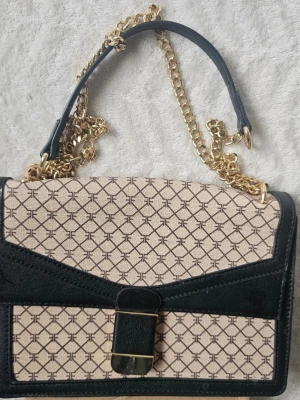 Mönstrad handväska från Even&Odd - Snygg handväska från Even&Odd med beige och svart geometriskt mönster, svarta detaljer i konstläder och guldfärgad kedjerem. Väskan har ett elegant spänne framtill och en stilren, rektangulär form. Perfekt accessoar för att lyfta din outfit.