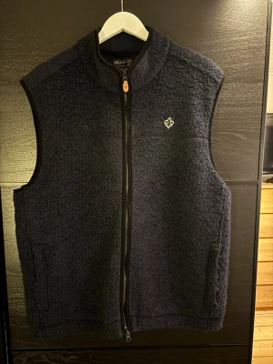 Mörkblå fleeceväst från Morris - Fet mörkblå fleeceväst från Morris med med dragkedja framtill. Västen har två fickor och en liten vit logga på bröstet. Perfekt för lager-på-lager och har en mjuk, teddy-liknande struktur. Hör gärna av dig om du har några frågor!