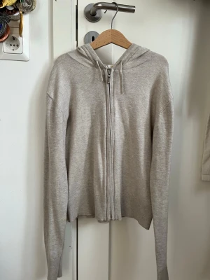 Beige zip-hoodie med huva - Snygg beige hoodie med dragkedja framtill och huva med dragsko. Modellen är croppad och har långa ärmar. Tillverkad i mjukt, stickat material som känns skönt mot huden. Perfekt för en avslappnad och trendig look.