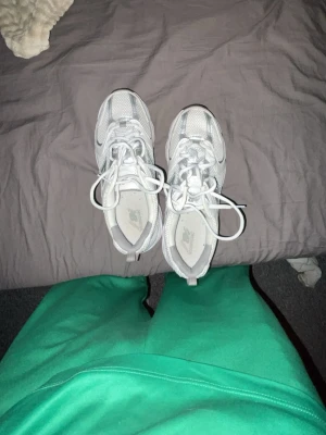 Vita New Balance sneakers - Säljer ett par fräscha vita New Balance sneakers med silvriga detaljer. Skorna har en chunky sula och är tillverkade i mesh och syntetmaterial för en sportig look. Klassisk N-logga på sidan och snörning framtill. Perfekta för dig som gillar streetwear.