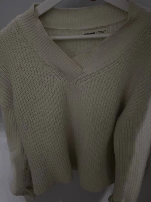 Beige stickad tröja med v-ringning - Mysig beige stickad tröja från Pull & Bear med bred v-ringning och ribbstickade detaljer. Tröjan har en avslappnad passform och långa ärmar, perfekt för lager på lager under kyliga dagar.