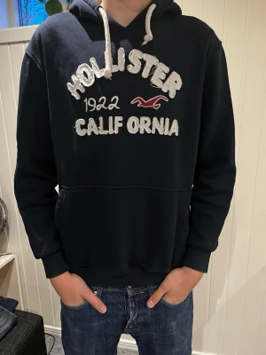 Svart Hollister hoodie med brodyr - Cool svart hoodie från Hollister med broderad text 'Hollister 1922 California' och logotyp på bröstet. Den har en stor huva med vita snören och en praktisk magficka. Perfekt för dig som gillar avslappnad stil och klassiska märken.